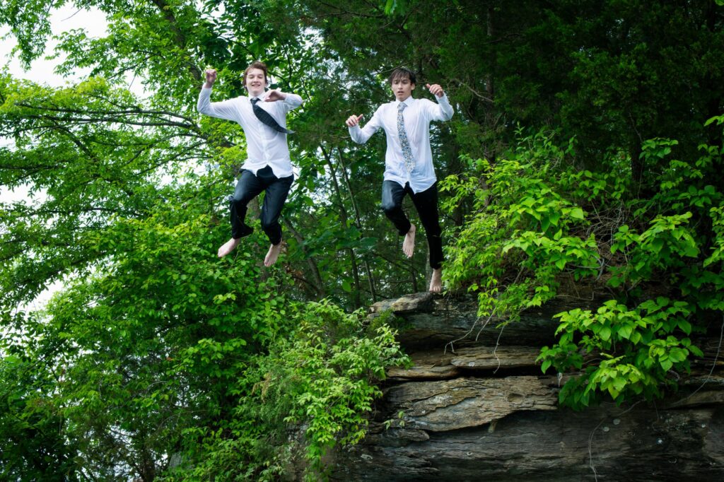 two senior boys jumping off a rocky cliff with green trees into Dale Hollow Lake destination portrait sessions senior unique portraits taken by Krista Nutter Photography