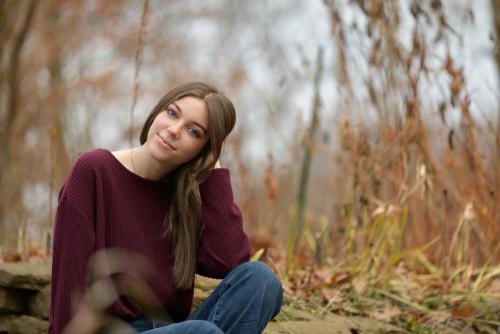 senior girl with brown hair in a burgundy shirt sitting in a field of tall grass in fall senior portrait by Krista Nutter Photography Cincinnati