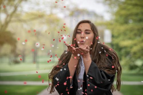 college senior girl with brown hair and a black graduation gown blowing red and white biodegradeable confetti on the Ohio State campus portrait by Krista Nutter Photography Cincinnati