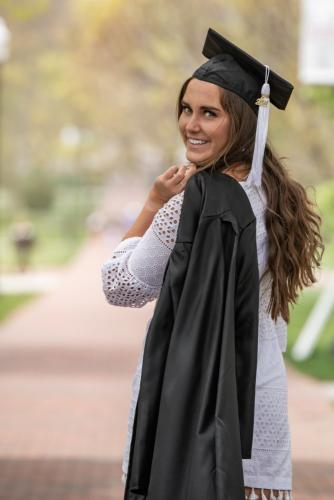 college senior girl in white dress with black graduation gown over shoulder looking backwards over her shoulder as she walks away from the camera on Ohio State campus senior portrait by Krista Nutter Photography Cincinnati