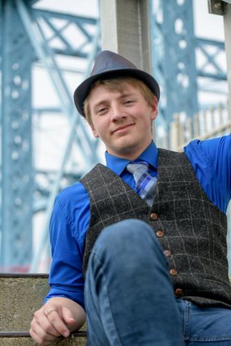 senior boy in blue shirt tie vest and hat sitting on stairs with the Roebling Suspension Bridge behind him senior portrait by Krista Nutter Photography Cincinnati