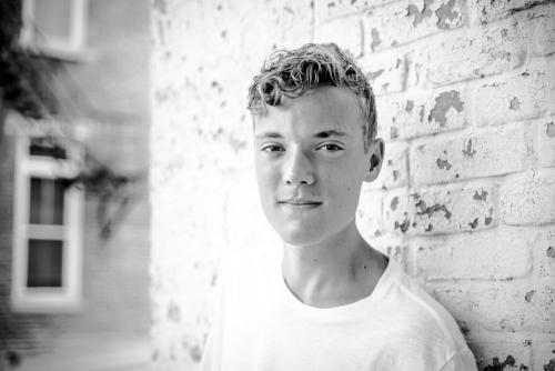 black and white image of a senior boy leaning against a white brick wall senior portrait by Krista Nutter Photography Cincinnati