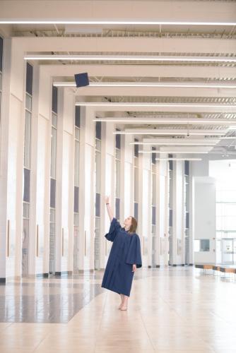 senior girl in blue cap and gown tossing her graduation cap in the air in a bright and airy indoor atrium space senior portrait by Krista Nutter Photography Cincinnati