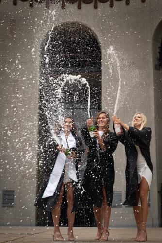 three college senior girls wearing black graduation gowns popping a bottle of champagne at the Ohio State Horseshoe Stadium rotunda senior portrait by Krista Nutter Photography Cincinnati