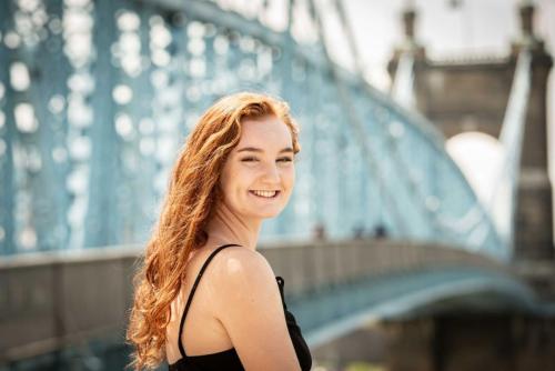senior girl with long red hair and black dress looking back over her shoulder with the blue Roebling Suspension bridge behind her senior portrait by Krista Nutter Photography Cincinnati