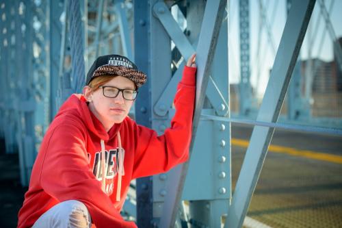 senior boy in red shirt kneeling casually on the blue Roebling Suspension Bridge senior portrait by Krista Nutter Photography Cincinnati