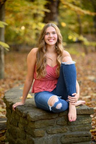 barefoot senior girl sitting on stone wall at the Cincinnati Nature Center with fall colors behind her senior portrait by Krista Nutter Photography Cincinnati