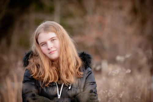 senior girl with blonde hair in a black coat with a fur hood standing in a field of tall grasses in winter at the Cincinnati Nature Center senior portrait by Krista Nutter Photography Cincinnati