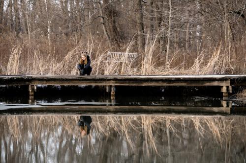 senior girl kneeling down next to water to view reflection and tall grasses behind her at the Cincinnati Nature Center winter senior portrait by Krista Nutter Photography Cincinnati
