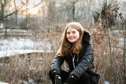 senior girl with blonde hair in a black coat with a fur hood sitting in a snowy field of tall grasses in winter at the Cincinnati Nature Center senior portrait by Krista Nutter Photography Cincinnati